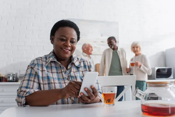 Smiling african american woman using smartphone near tea and blurred people in nursing home — Stock Photo