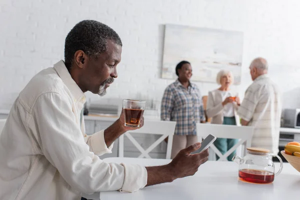 Sonriente hombre afroamericano mayor con té usando teléfono inteligente en un hogar de ancianos - foto de stock