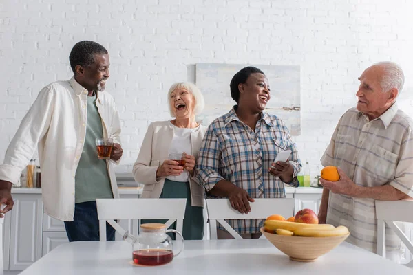 Des personnes multiethniques gaies tenant du thé et un smartphone tout en parlant dans une maison de retraite — Photo de stock