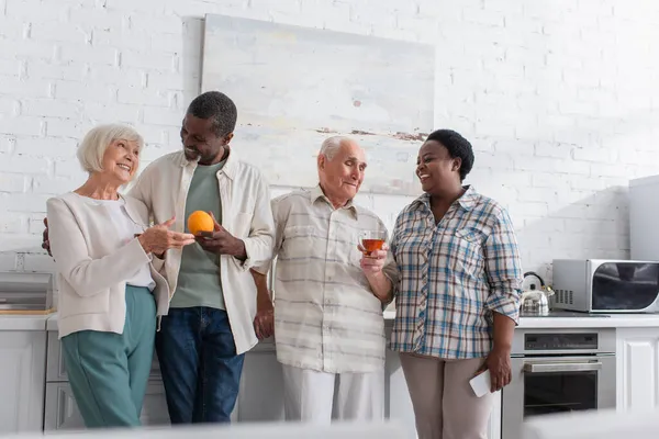Позитивні міжрасові літні люди з чаєм, фруктами та смартфонами розмовляють в будинку престарілих — стокове фото