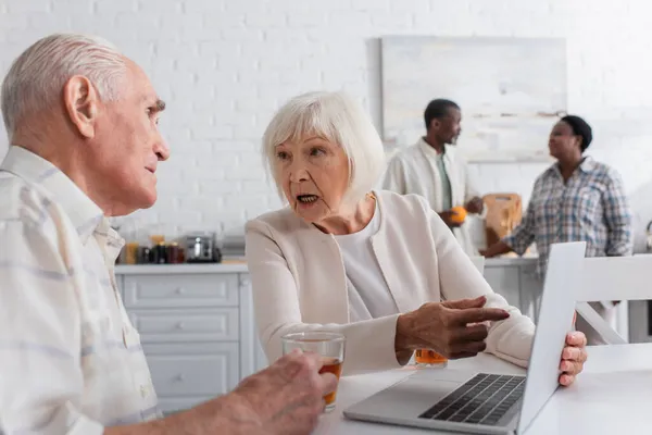 Mujer anciana señalando a la computadora portátil cerca de amigo mayor y té en el hogar de ancianos - foto de stock