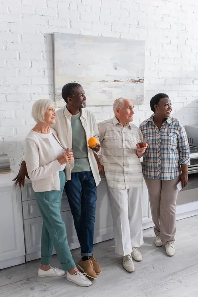 Personas mayores multiétnicas con té y teléfono inteligente de pie en la cocina en el hogar de ancianos — Stock Photo