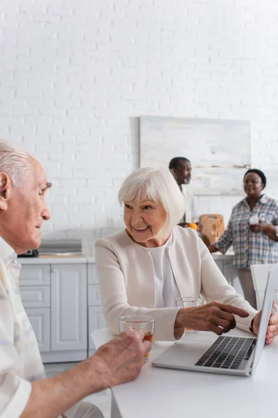 Mujer anciana sonriente apuntando a la computadora portátil cerca de amigo con té en el hogar de ancianos - foto de stock