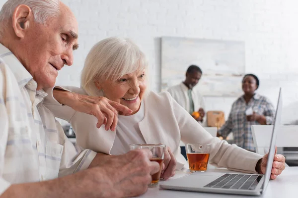 Положительная пожилая женщина указывает на ноутбук рядом с другом с чашкой чая в доме престарелых — стоковое фото