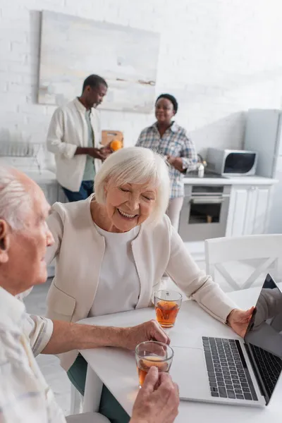 Веселая женщина сидит рядом со старшим другом с чаем и ноутбуком в доме престарелых — стоковое фото