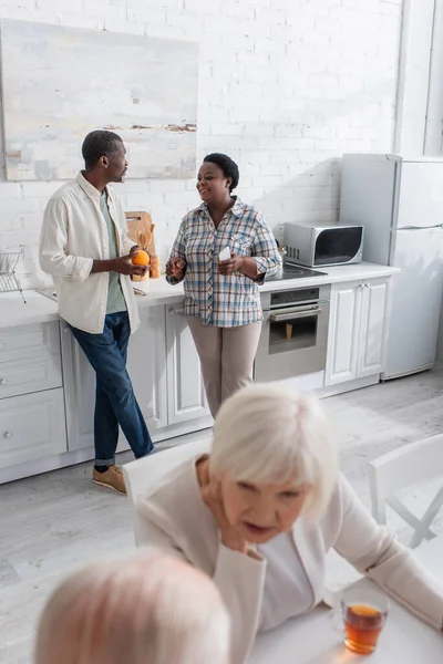 Sonrientes afroamericanos con fruta y smartphone hablando en cocina de hogar de ancianos - foto de stock