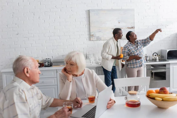 Afroamerikanerin mit Smartphone zeigt mit Finger auf Freundin mit Obst im Pflegeheim — Stockfoto