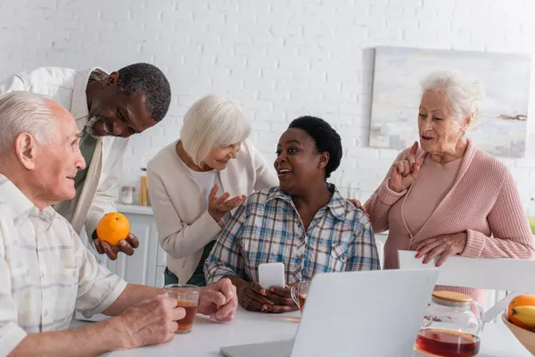 Positive interracial senior friends talking near tea and devices in nursing home — Stock Photo