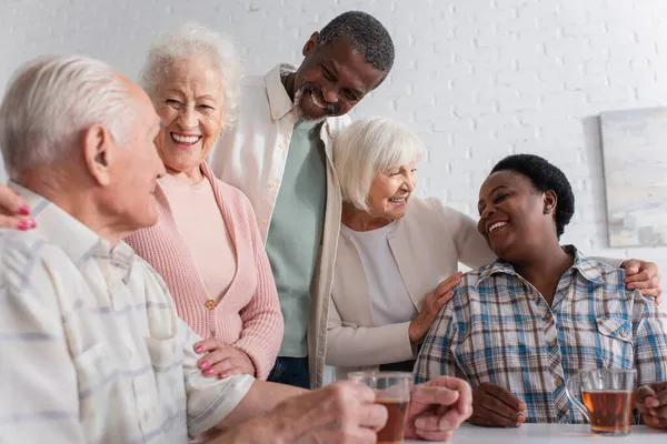 Positive interracial friends talking near tea in nursing home — Stock Photo