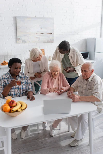 Позитивні багатоетнічні літні люди використовують ноутбук біля чаю та фруктів на кухні — стокове фото
