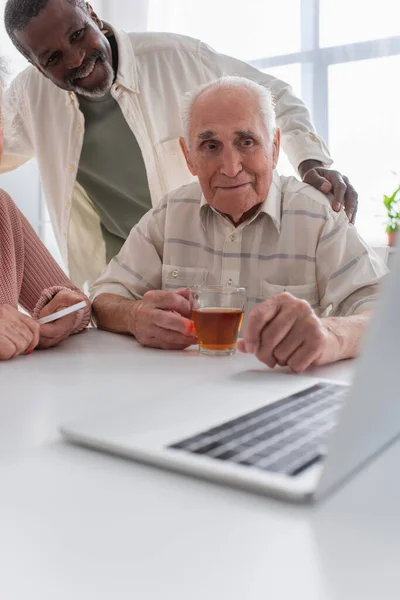 Lächelnder afrikanisch-amerikanischer Mann umarmt ältere Freunde mit Tee in der Nähe von Laptop im Pflegeheim — Stockfoto