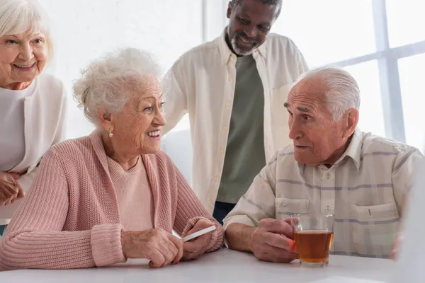 Amigos mayores multiétnicos positivos con teléfono inteligente y té hablando en el hogar de ancianos - foto de stock