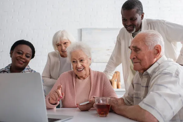 Mujer mayor sonriente sosteniendo el teléfono inteligente y apuntando a la computadora portátil cerca de amigos interracial en el hogar de ancianos - foto de stock