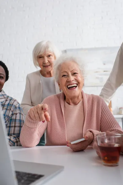 Mujer mayor alegre con teléfono inteligente apuntando a la computadora portátil cerca del té y amigos interracial en el hogar de ancianos - foto de stock