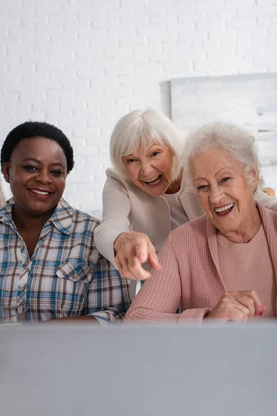 Mulher sênior feliz apontando para laptop perto de amigos inter-raciais em casa de repouso — Fotografia de Stock