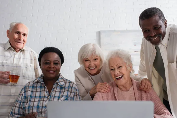 Multiethnische Senioren mit Tee schauen im Pflegeheim auf Laptop — Stockfoto