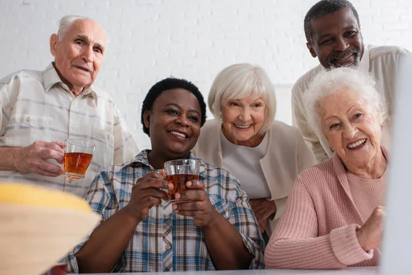 Cheerful multiethnic senior people holding cups of tea near blurred laptop in nursing home — Stock Photo