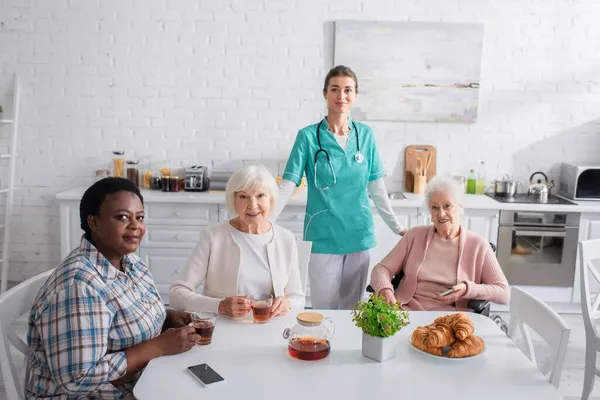 Enfermera joven de pie cerca de las mujeres mayores interracial con dispositivos y té en el hogar de ancianos - foto de stock