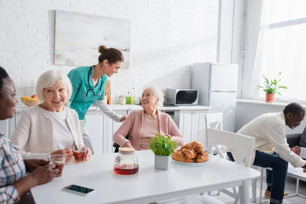 Nurse talking to elderly woman in wheelchair near interracial people in nursing home — Stock Photo