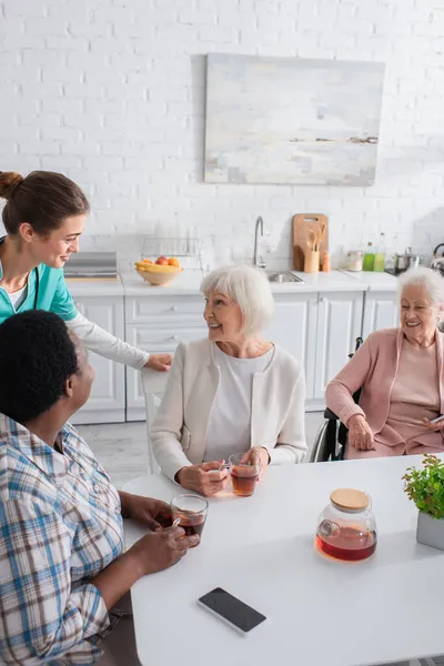 Lächelnde multiethnische ältere Frauen im Gespräch mit Krankenschwester in der Nähe von Tee im Pflegeheim — Stockfoto