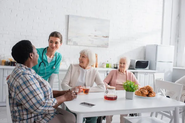 Krankenschwester in Uniform spricht mit interrassischen Patienten in der Nähe von Smartphone und Tee im Pflegeheim — Stockfoto