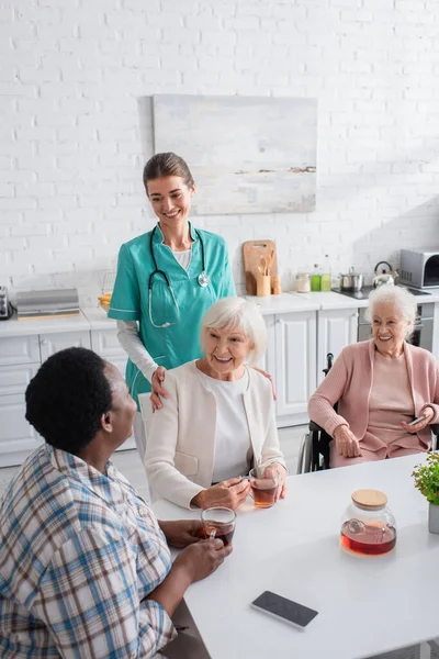 Infirmière souriante debout près de patients multiethniques avec smartphones et thé dans la cuisine de la maison de soins infirmiers — Photo de stock