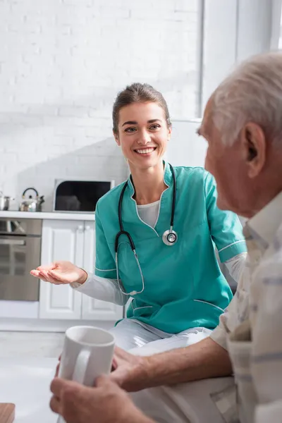 Lächelnde Krankenschwester zeigt mit Hand auf verschwommenen Senior mit Tasse im Pflegeheim — Stockfoto