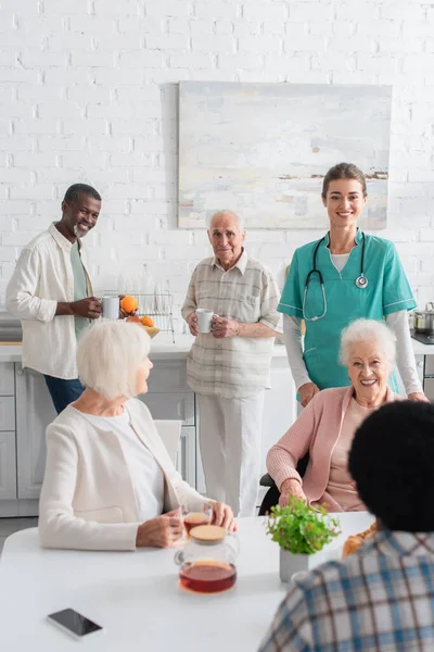 Улыбающиеся межрасовые люди с чаем разговаривают рядом с молодой медсестрой в доме престарелых — стоковое фото