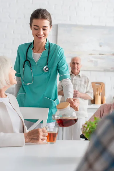 Улыбающаяся медсестра держит чайник рядом с улыбающимися пожилыми пациентами в доме престарелых — стоковое фото