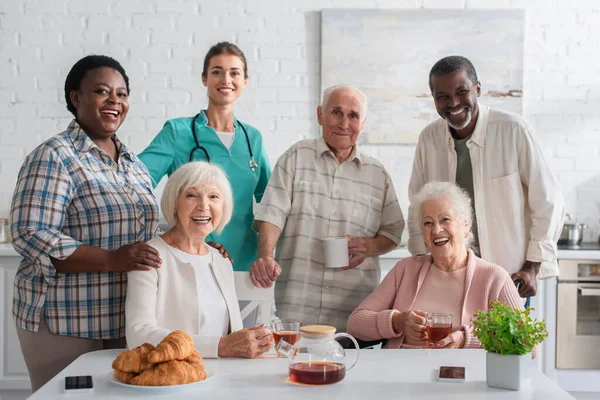 Smiling nurse standing near interracial patients and tea in nursing home — Stock Photo