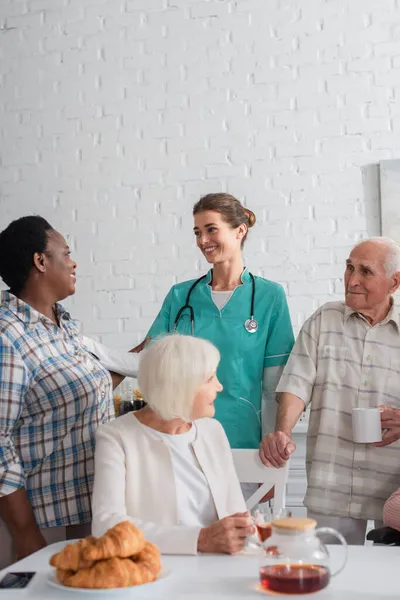 Fröhliche Krankenschwester im Gespräch mit interrassischen Patienten bei Croissants und Tee im Pflegeheim — Stockfoto