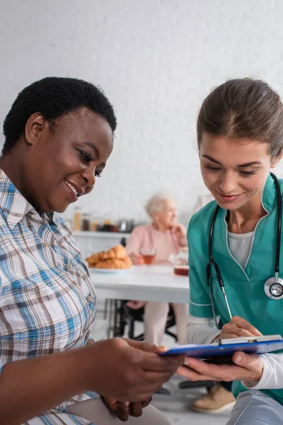 Lächelnde Krankenschwester und afrikanisch-amerikanische Patientin beim Blick auf Klemmbrett im Pflegeheim — Stockfoto