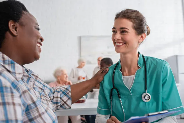 Sorrindo afro-americana olhando para jovem enfermeira com prancheta em lar de idosos — Fotografia de Stock