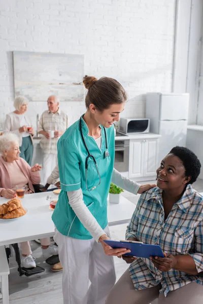 Mujer afroamericana sosteniendo portapapeles cerca de enfermera sonriente en hogar de ancianos - foto de stock