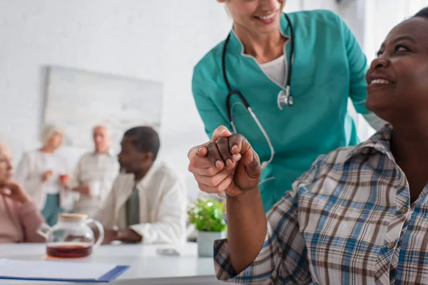 Verschwommene Krankenschwester hält Hand von positiver afrikanisch-amerikanischer Patientin in der Nähe von Menschen im Pflegeheim — Stockfoto