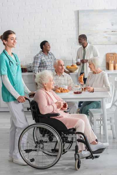 Enfermera sonriente parada cerca de una mujer mayor en silla de ruedas sosteniendo té en un hogar de ancianos - foto de stock