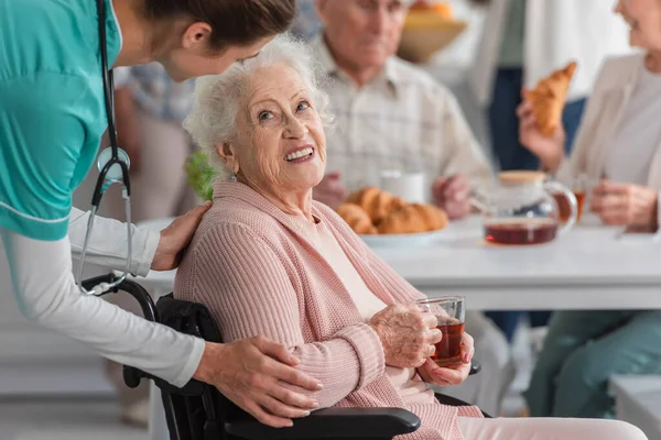 Infermiera che abbraccia sorridente donna anziana con tè in sedia a rotelle in casa di cura — Foto stock