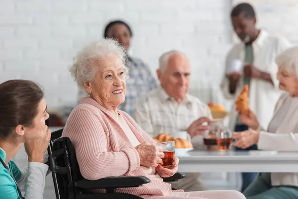 Senior woman holding tea in wheelchair near nurse and blurred interracial friends in nursing home — Stock Photo