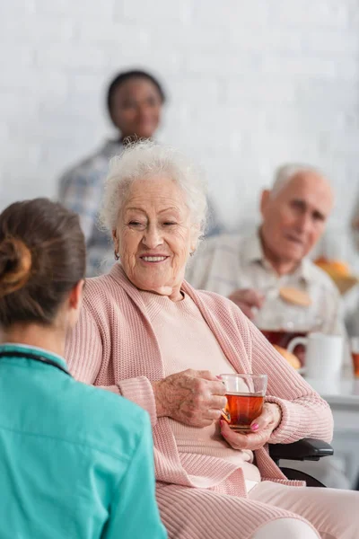 Mulher idosa sorridente em cadeira de rodas segurando xícara de chá perto de enfermeira turva em casa de repouso — Fotografia de Stock