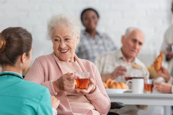 Positive senior woman holding tea while talking to blurred nurse in nursing home — Stock Photo