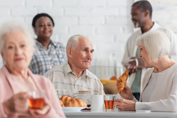 Mujer mayor sonriente sosteniendo croissant y té cerca de amigo en el hogar de ancianos - foto de stock