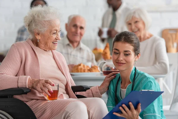 Улыбающаяся пожилая женщина с чаем в инвалидной коляске рядом с медсестрой с планшетом в доме престарелых — стоковое фото
