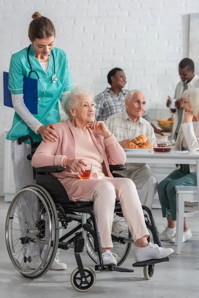 Enfermera con portapapeles de pie cerca del paciente con té en silla de ruedas en el hogar de ancianos - foto de stock