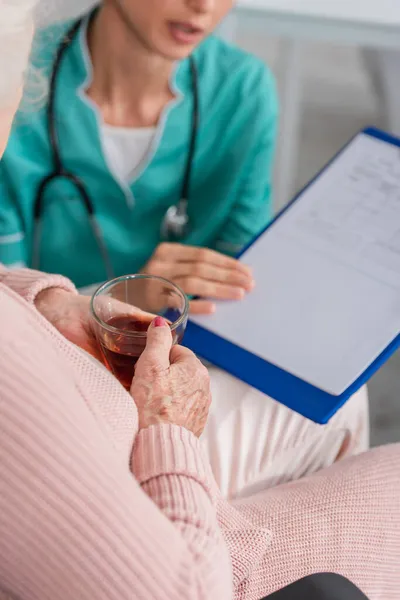 Ausgeschnittene Ansicht einer Seniorin mit Tee in der Nähe einer verschwommenen Krankenschwester mit Klemmbrett im Pflegeheim — Stockfoto