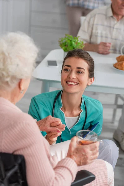 Infermiera sorridente che tiene la mano della donna anziana con tè in sedia a rotelle nella casa di cura — Foto stock