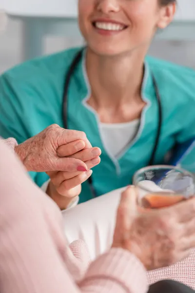 Vista recortada de la enfermera sonriente cogida de la mano del paciente mayor con té en el hogar de ancianos - foto de stock