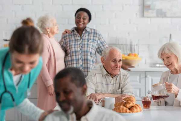 Mujer mayor sosteniendo tetera cerca de amigo y borrosa gente interracial en el hogar de ancianos - foto de stock