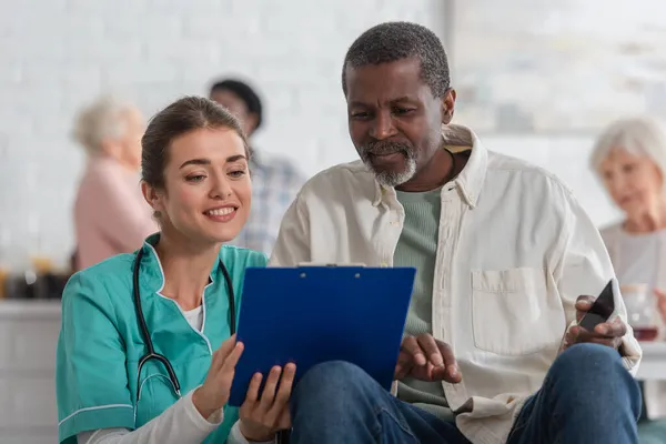 Smiling nurse holding clipboard near aftican american patient with smartphone in nursing home — Stock Photo