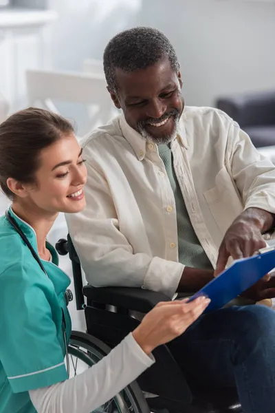 Усміхнена молода медсестра тримає кишеню біля афроамериканського пацієнта в інвалідному візку в будинку престарілих — стокове фото