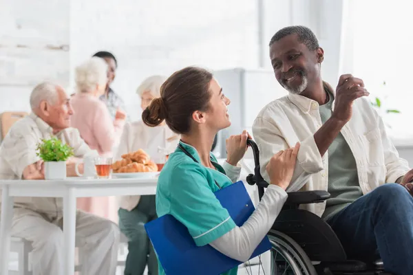 Enfermera alegre con portapapeles hablando con hombre afroamericano en silla de ruedas en asilo de ancianos - foto de stock